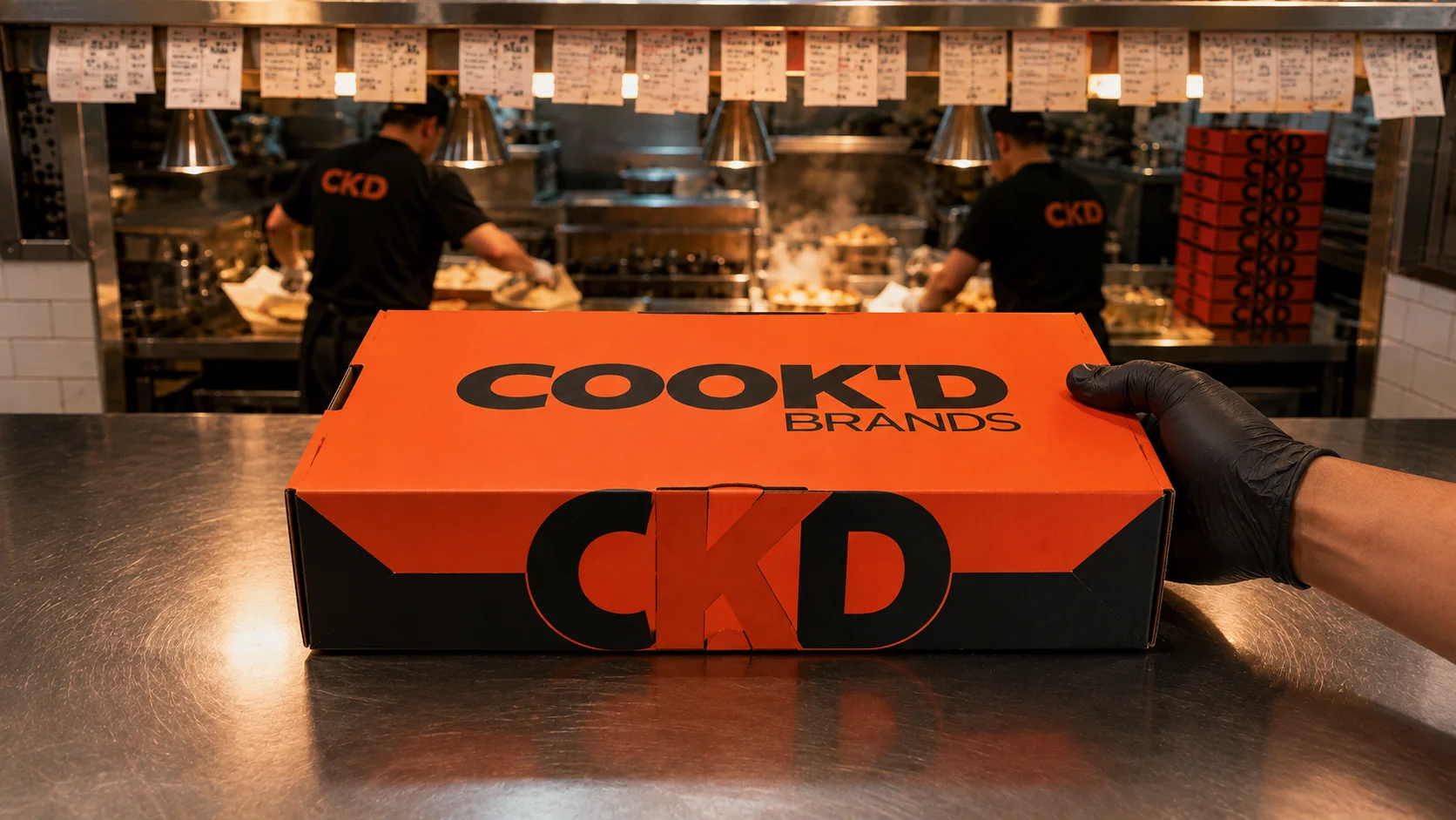 The closed Cook'd Lunchbox on the pass, with a busy kitchen behind it